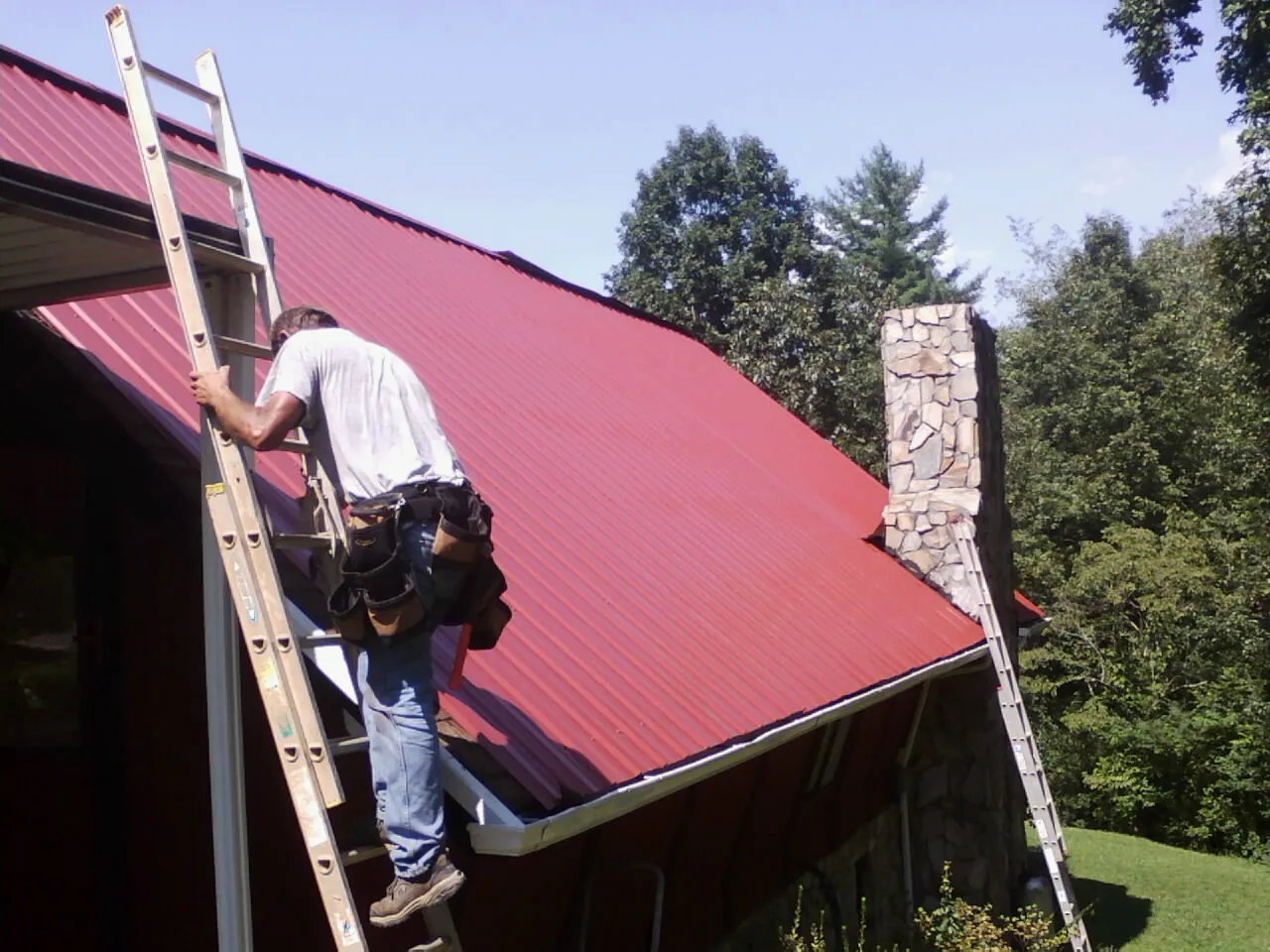 Roofer on a ladder beside a red metal roof and stone chimney — Soffit & Fascia Repair by Roofing Brothers in Palmer Lake